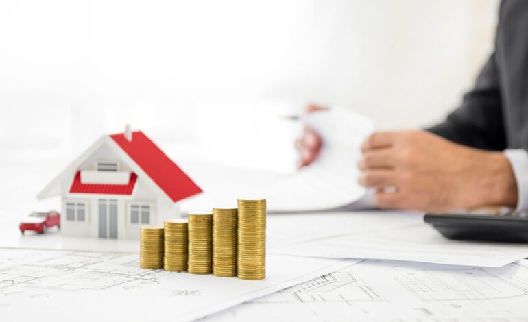 Pieniądze i model domu na papierze projektowym przy stole z rozmytym biznesmenem w tle - koncepcja finansowa nieruchomości i nieruchomości