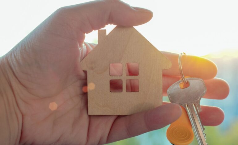 W ręku mężczyzny mały drewniany domek i klucz, na tle słońca w letni dzień. Koncepcja - kupowanie nieruchomości, marzenie o własnym domu, niedrogie mieszkania, sprzedaż nieruchomości.