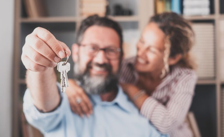 Nacisk na klucze, trzymane przez podekscytowanych młodych małżonków właścicieli domów. Szczęśliwa para małżeńska świętująca przeprowadzkę do nowego domu, demonstrująca klucze, stojąca w mieszkaniu, koncepcja kredytu hipotecznego na nieruchomości.